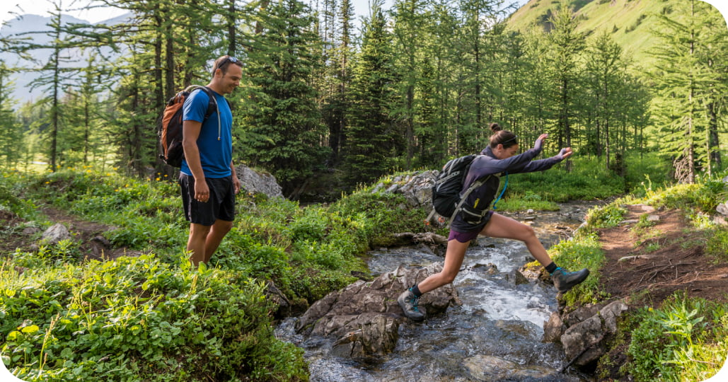 Two hikers jumping over a small creek one at a time in the middle of a forest