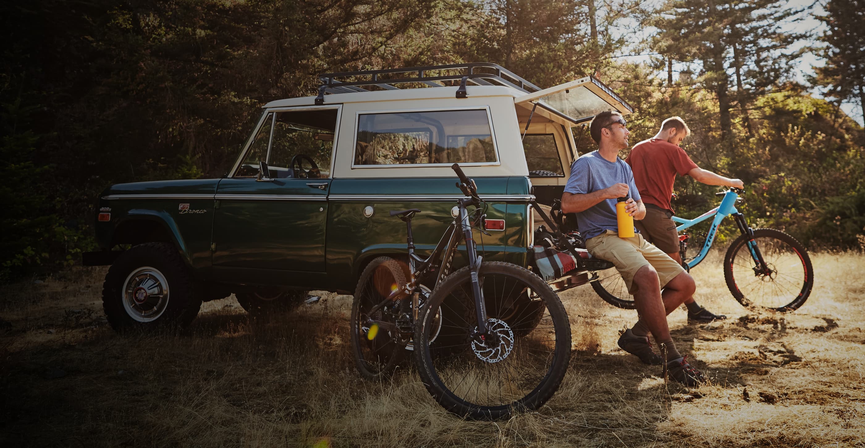 A biker with short brown hair and sunglasses sits on the tailgate of a green and white Ford Bronco opening a yellow water bottle, and another biker with short blond hair stands next to them inspecting a bright blue mountain bike. The vehicle is parked in a grassy field surrounded by trees and a black mountain bike leans against its side.